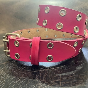 May include: A bright pink leather belt with silver grommets and a silver buckle. The belt is rolled up, showcasing the multiple holes for adjustment. The leather has a smooth texture.