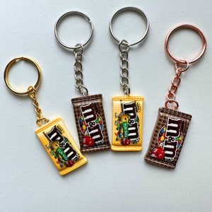Op de afbeelding: Vier sleutelhangers met miniatuur replica's van M&M's candybars. Twee zijn bruin, twee zijn geel, met het M&M's logo en de tekst "Fun Size". Elke sleutelhanger heeft een andere metalen afwerking: goud, zilver en ros&eacute;goud.