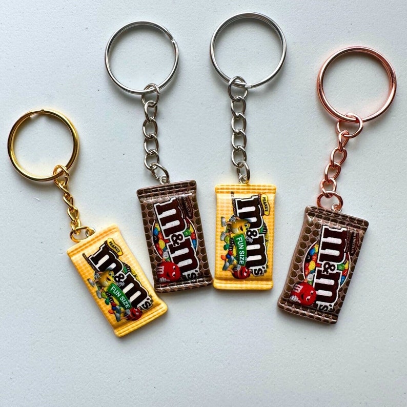 Op de afbeelding: Vier sleutelhangers met miniatuur replica's van M&M's candybars. Twee zijn bruin, twee zijn geel, met het M&M's logo en de tekst "Fun Size". Elke sleutelhanger heeft een andere metalen afwerking: goud, zilver en ros&eacute;goud.