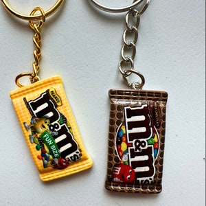 Op de afbeelding: Twee sleutelhangers met miniatuur snoepreepontwerpen. E&eacute;n is geel met de tekst "Peanut" en "Fun Size", de andere is bruin. Beide hebben metalen sleutelringen en kettingen.
