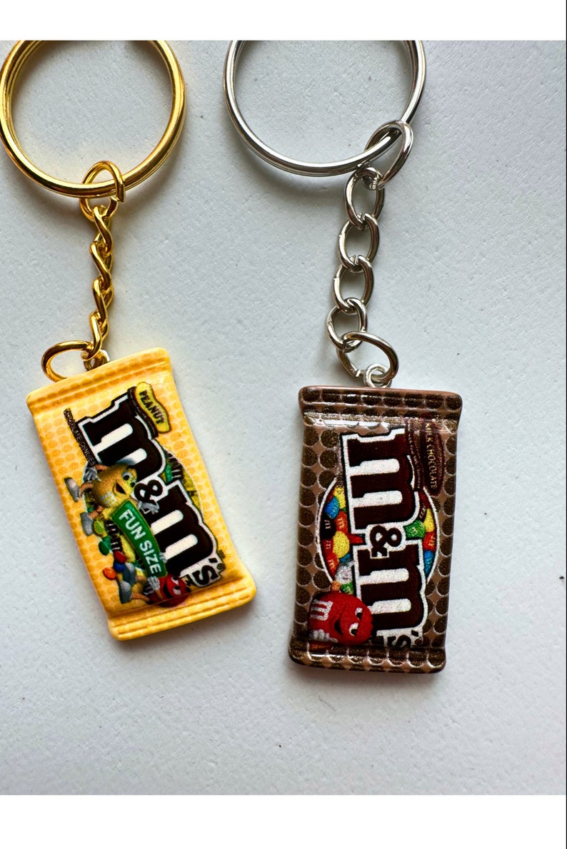Op de afbeelding: Twee sleutelhangers met miniatuur snoepreepontwerpen. E&eacute;n is geel met de tekst "Peanut" en "Fun Size", de andere is bruin. Beide hebben metalen sleutelringen en kettingen.