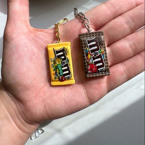 Op de afbeelding: Twee miniatuur M&M's candy bar sleutelhangers. E&eacute;n is geel met de tekst "Fun Size" en de andere is bruin. Beide hebben zilveren sleutelringen en goudkleurige kettingen. De sleutelhangers worden in de open handpalm van een persoon gehouden.