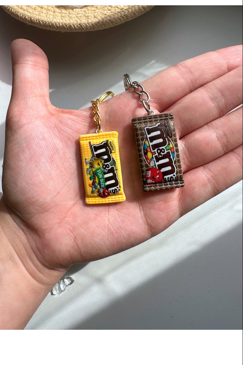 Op de afbeelding: Twee miniatuur M&M's candy bar sleutelhangers. E&eacute;n is geel met de tekst "Fun Size" en de andere is bruin. Beide hebben zilveren sleutelringen en goudkleurige kettingen. De sleutelhangers worden in de open handpalm van een persoon gehouden.