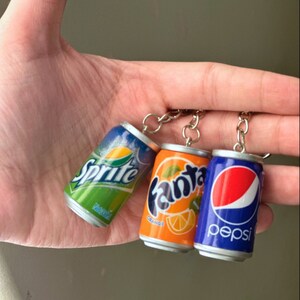 May include: Three miniature soda can keychains. The cans are silver with green, orange, and blue labels. The labels read "Sprite", "Fanta Orange", and "Pepsi".