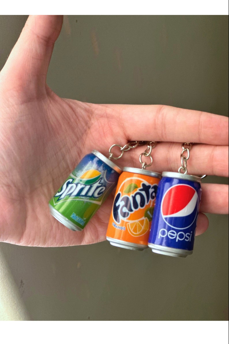 May include: Three miniature soda can keychains. The cans are silver with green, orange, and blue labels. The labels read "Sprite", "Fanta Orange", and "Pepsi".