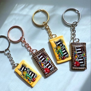 Op de afbeelding: Vier sleutelhangers met miniatuur replica's van M&M's candybars. Elke sleutelhanger heeft een verschillend gekleurde metalen ring en ketting. De candybar replica's zijn geel en bruin, met het M&M's logo en tekst.
