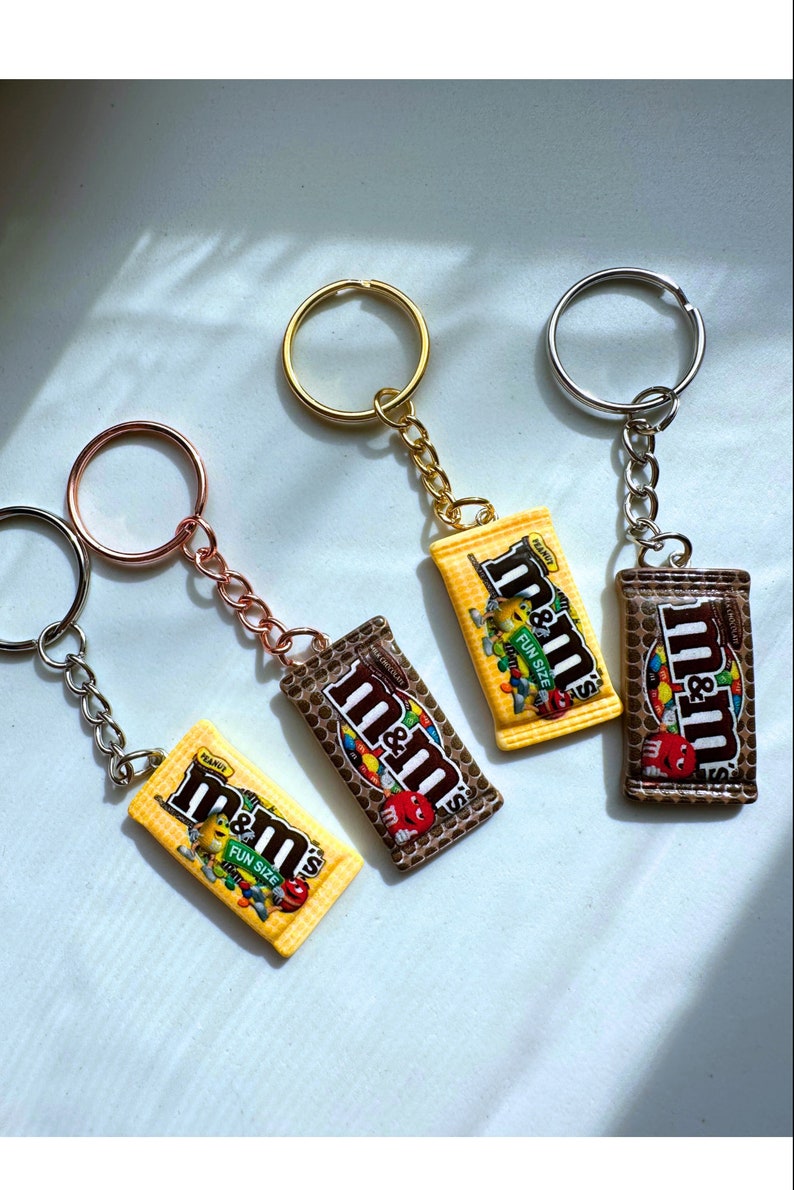 Op de afbeelding: Vier sleutelhangers met miniatuur replica's van M&M's candybars. Elke sleutelhanger heeft een verschillend gekleurde metalen ring en ketting. De candybar replica's zijn geel en bruin, met het M&M's logo en tekst.