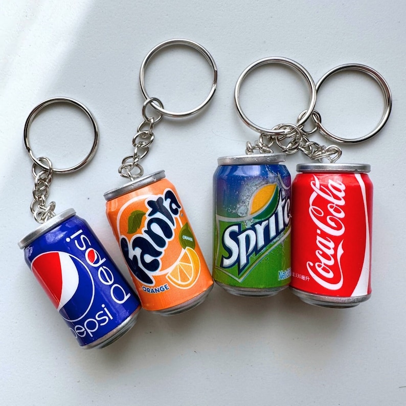 May include: Four miniature soda can keychains, each featuring a different brand: Pepsi, Fanta Orange, Sprite, and Coca-Cola. The keychains are attached to silver key rings.