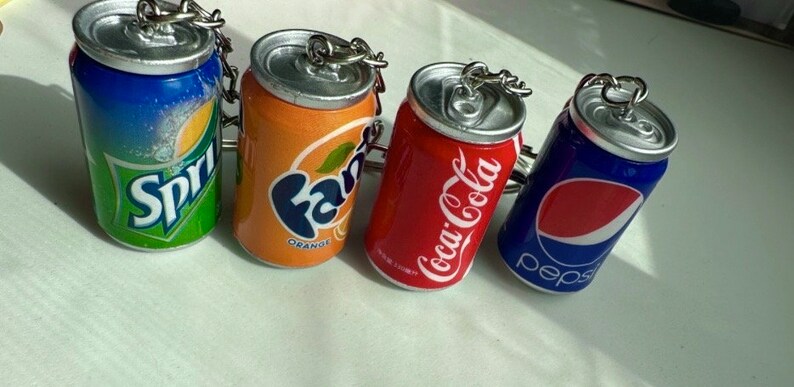May include: Four miniature soda can keychains. The cans are blue, orange, red, and blue with the logos "Sprite", "Fanta Orange", "Coca-Cola", and "Pepsi" respectively.