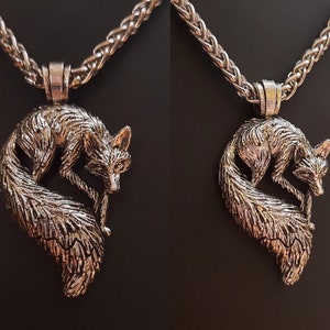 Puede incluir: Un collar de plata con un colgante de zorro. El zorro est&aacute; enroscado con la cola envuelta alrededor de su cuerpo. El collar est&aacute; sobre un fondo negro.