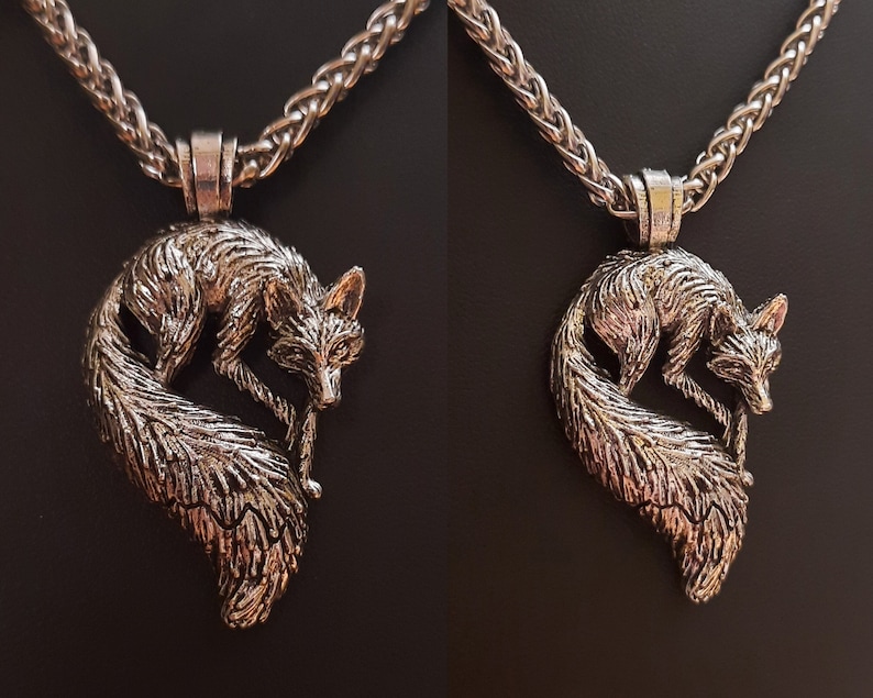 Puede incluir: Un collar de plata con un colgante de zorro. El zorro est&aacute; enroscado con la cola envuelta alrededor de su cuerpo. El collar est&aacute; sobre un fondo negro.