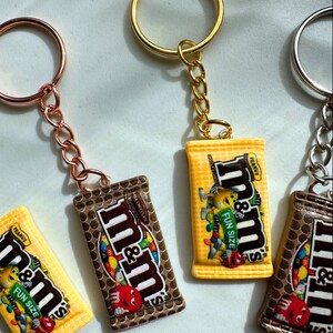 Op de afbeelding: Sleutelhangers met miniatuur replica's van M&M's chocoladerepen. De sleutelhangers hebben een gouden, zilveren en ros&eacute;gouden ring en ketting. De chocoladereep replica's zijn bruin en geel met het M&M's logo en de woorden "FUN SIZE" en "PEANUT".