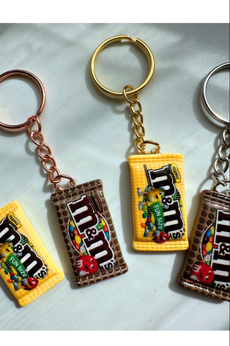 Op de afbeelding: Sleutelhangers met miniatuur replica's van M&M's chocoladerepen. De sleutelhangers hebben een gouden, zilveren en ros&eacute;gouden ring en ketting. De chocoladereep replica's zijn bruin en geel met het M&M's logo en de woorden "FUN SIZE" en "PEANUT".