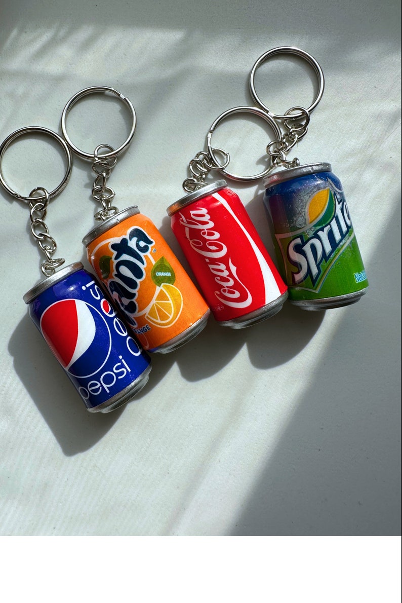 May include: Four miniature soda can keychains, featuring the brands Pepsi, Fanta, Coca-Cola, and Sprite. The keychains are silver with a metallic finish.