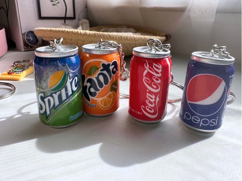May include: Four miniature soda can keychains, each featuring a different brand: Sprite, Fanta Orange, Coca-Cola, and Pepsi. The keychains are silver and have a realistic can design.