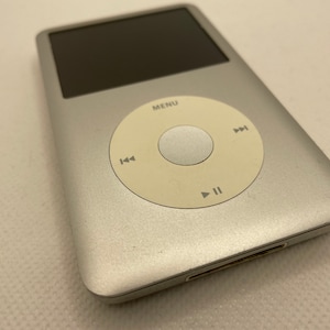 May include: A silver Apple iPod with a white click wheel and a black screen. The iPod has buttons for play, pause, fast forward, and rewind. The text "MENU" is visible on the click wheel.