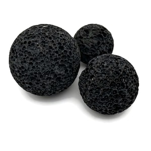 Può includere: Tre sfere di roccia lavica nera di diverse dimensioni sono disposte su uno sfondo bianco. La consistenza porosa delle rocce è chiaramente visibile. Queste sfere decorative sono spesso utilizzate in aromaterapia o come elementi decorativi.