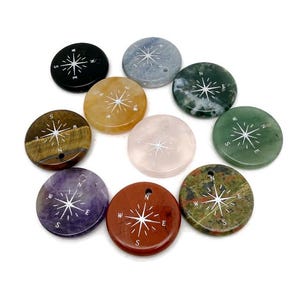 Könnte beinhalten: Eine Sammlung runder, polierter Edelsteinanhänger, die jeweils ein weißes Kompassdesign aufweisen. Die Anhänger variieren in der Farbe, darunter Schwarz, Blau, Gelb, Braun, Lila, Rot, Rosa, Grün und ein gesprenkeltes Grün und Braun. Jeder Anhänger hat ein kleines Loch.