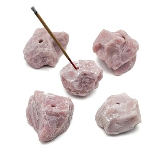 May include: Five rough-cut, light pink stone incense holders. One holder has a lit incense stick. The holders are irregularly shaped with a hole drilled in the top to hold incense. The background is white.