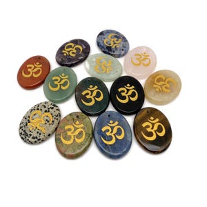 Puede incluir: Una colección de colgantes de piedras preciosas de forma ovalada en varios colores, incluyendo rojo, verde, negro y azul. Cada colgante presenta un símbolo Om dorado. Los colgantes están dispuestos sobre una superficie blanca.