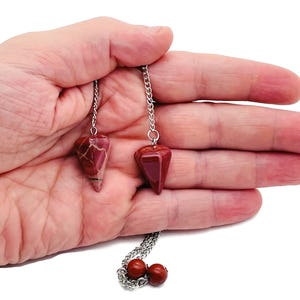 Puede incluir: Dos péndulos de piedras preciosas rojas y marrones con cadenas plateadas. Los péndulos se sostienen en una mano sobre un fondo blanco. Las piedras tienen forma triangular.