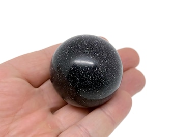 May include: A black sphere with a speckled texture, resembling a miniature planet. The sphere is smooth and polished, with a slight sheen.