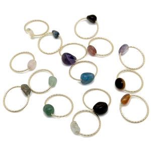 Puede incluir: Una colección de delicados anillos dorados, cada uno con una gema de diferente color. Las gemas incluyen tonos de púrpura, azul, verde, marrón y transparente. Los anillos están dispuestos sobre una superficie blanca.