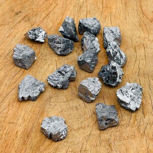 May include: A collection of 15 silver-colored, rough-hewn, metallic mineral stones. The stones are various sizes and shapes, and are arranged on a wooden surface.