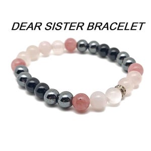 May include: A bracelet made with pink, rose quartz, and black hematite beads. The bracelet has the words "Dear Sister Bracelet" printed above it.