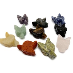 Può includere: Una collezione di figurine a forma di testa di lupo scolpite in pietra di vari colori, tra cui giallo, verde, rosso e viola. Le figurine sono esposte su una superficie bianca.