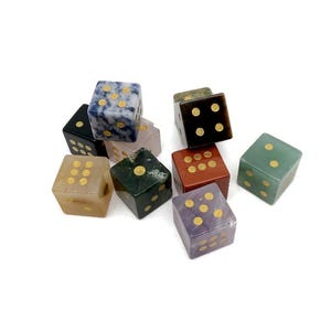 May include: A collection of gemstone dice in various colors, including blue, green, red, and purple. Each die features gold-colored dots. The dice are arranged on a white background.