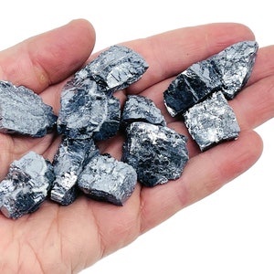 May include: A collection of raw, metallic gray, irregularly shaped, and rough-textured,  crystals. The crystals are in a hand, and the background is white.