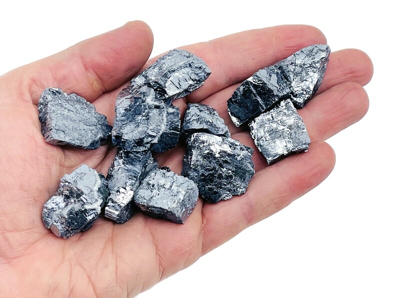 May include: A collection of raw, metallic gray, irregularly shaped, and rough-textured,  crystals. The crystals are in a hand, and the background is white.
