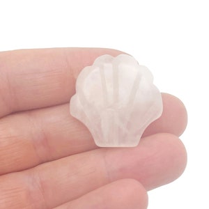May include: A carved, light pink seashell figurine. The seashell is held in a hand against a white background. The seashell has a detailed, ridged design and a smooth, polished finish. The figurine is a decorative item.