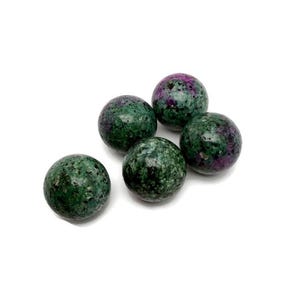 Könnte beinhalten: Fünf runde Edelsteine mit gesprenkeltem grün-violettem Aussehen. Die Steine sind poliert und glatt, mit einem natürlichen, organischen Aussehen. Die Edelsteine liegen auf einer weißen Oberfläche.
