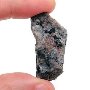 May include: A small, rough, gray and black rock with a slightly pink hue. The rock has a jagged, irregular shape.
