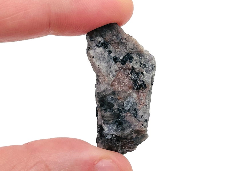 May include: A small, rough, gray and black rock with a slightly pink hue. The rock has a jagged, irregular shape.