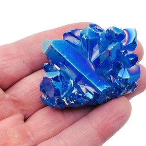 Puede incluir: Un cúmulo de cristales de cuarzo azul iridiscente, también conocidos como cuarzo aura, con un brillo metálico. Los cristales están dispuestos en un cúmulo y tienen una variedad de formas y tamaños.