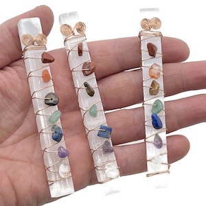 Może przedstawiać: Trzy różdżki selenitowe owinięte miedzianym drutem z siedmioma kamieniami czakr. Kamienie są czerwone, pomarańczowe, żółte, zielone, niebieskie, indygo i fioletowe.