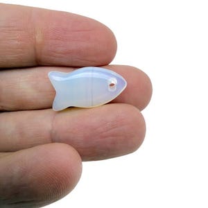 May include: A small, translucent, opalescent fish-shaped bead. The bead has a hole through it, and is held in a person's hand. The fish has a rounded body and a small tail.