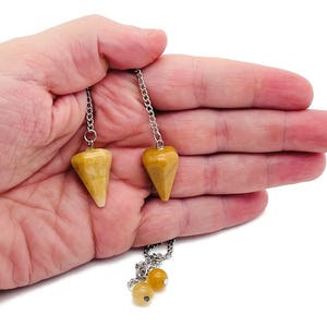 May include: Two yellow-brown gemstone pendulums with silver chains and a cluster of yellow beads. The pendulums are held in an open hand against a white background. The pendulums have a cone shape.