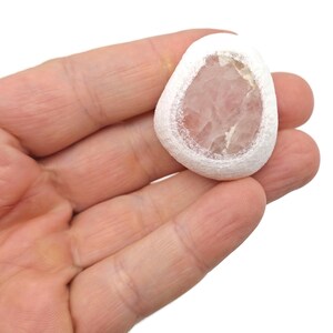 May include: A white, irregularly shaped stone with a light pink, translucent center. The stone is held in a hand against a white background. The stone has a textured, porous appearance.