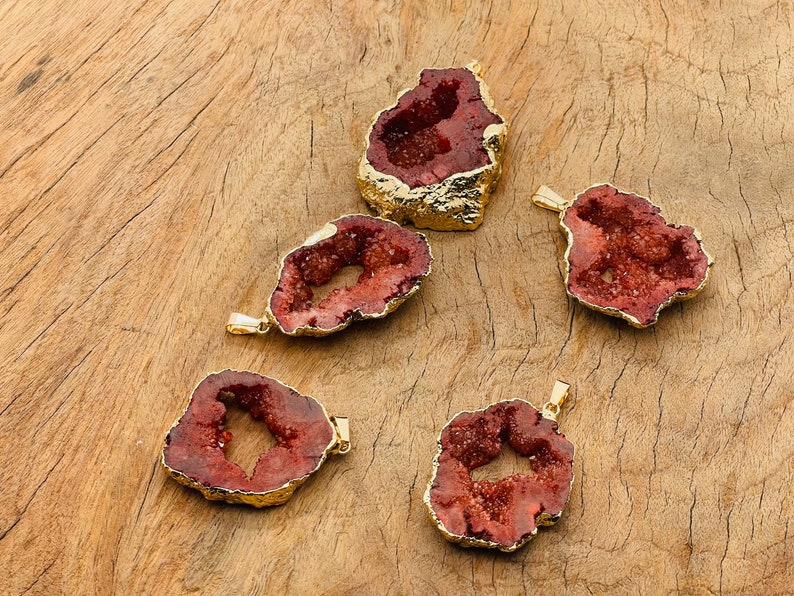 May include: Five red agate slices with gold-plated edges. Each slice has a natural, irregular shape and a hollow center. The slices are arranged on a wooden surface.
