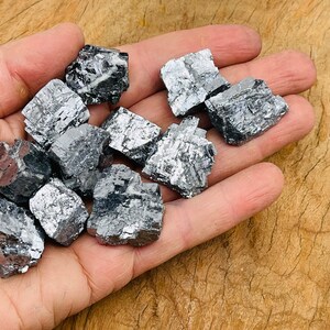 May include: A collection of raw, metallic gray pyrite crystals in various sizes and shapes. The crystals have a rough, textured surface and are arranged in a hand.