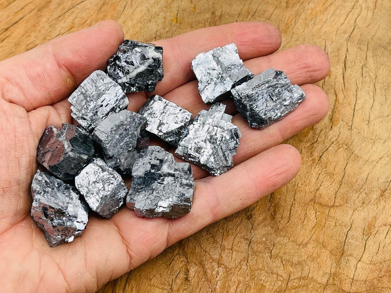 May include: A collection of raw, metallic gray pyrite crystals in various sizes and shapes. The crystals have a rough, textured surface and are arranged in a hand.