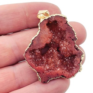 Puede incluir: Un colgante de geoda de ágata roja con un borde chapado en oro. La geoda está cortada por la mitad para revelar las formaciones cristalinas en el interior.