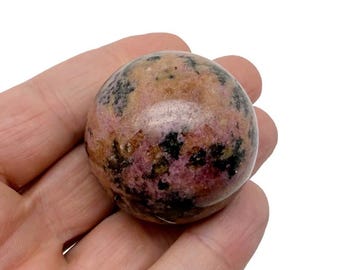 May include: A polished, spherical gemstone with a mix of colors. The stone features a base of pink and tan hues, with darker black and brown spots scattered across the surface. The gemstone is held in a hand.