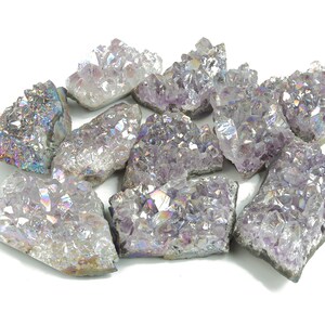 Puede incluir: Un conjunto de cristales de amatista con una superficie brillante e iridiscente. Los cristales, de varios tamaños y formas, muestran tonos de púrpura y destellos de arcoíris. Están dispuestos sobre un fondo blanco.