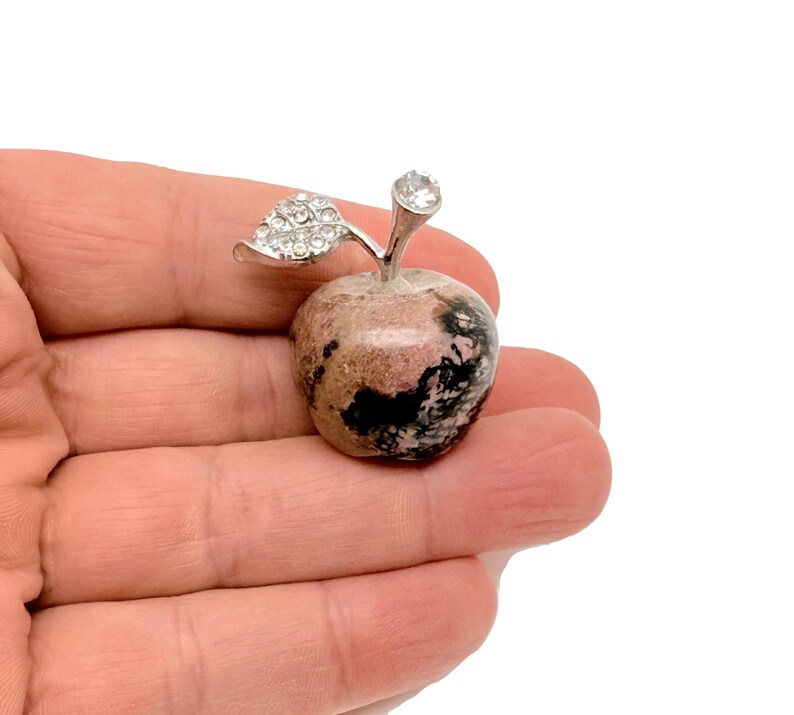 May include: A pink and black gemstone apple with a silver leaf and a clear rhinestone on top.