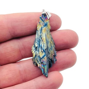 Puede incluir: Colgante con un cristal de cianita natural e iridiscente. El cristal muestra una mezcla de tonos azules, dorados y plateados, con una apariencia texturizada y en capas. Tiene una fianza plateada para colgar.